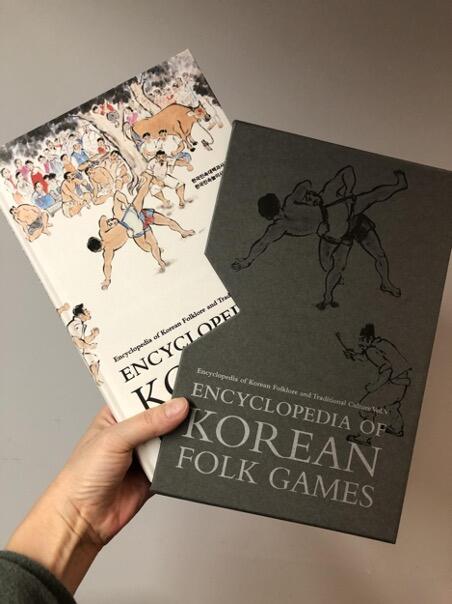 Image of hand holding Encyclopedia of Korean Folk Games hardcover volume and slipcase. 