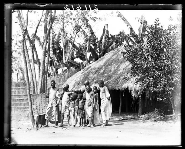 Children standing outside a school near Kafu River, a man holding a chart with Roman letters, Uganda, 1909-1911.