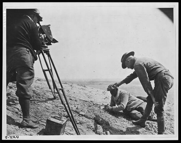 James B. Shackelford stands and films as George Olsen lies on the floor marking an object and Roy Chapman Andrews stands and points at Olsen.
