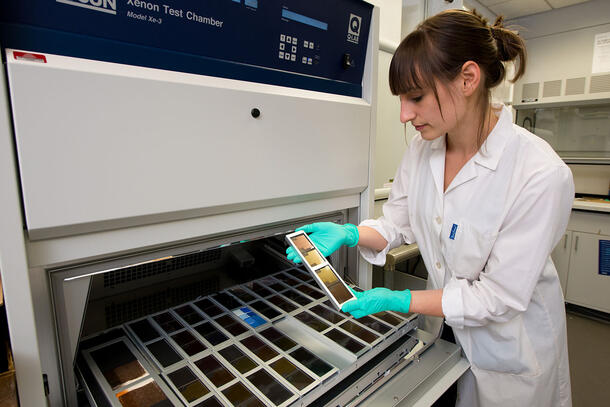 A woman in a lab coat and green gloves holding a dye sample in front of a tray of further samples. The tray is in the chamber of a large machine.