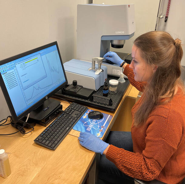 A woman sits at a desk in front of a computer. The screen shows colored spectra. She operates an FTIR spectrometer to her right.