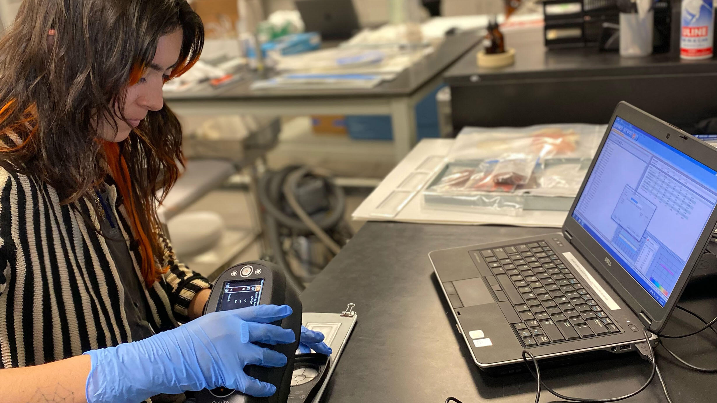 Scientist seated at a lab table holds a meter over a specimen and views the results on a laptop computer.