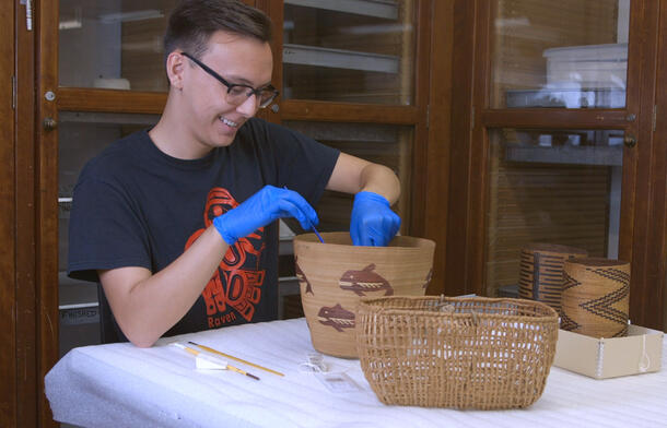 Brandon Castle is seated at a table, and wears gloves and uses small hand tools to repair a woven basket.