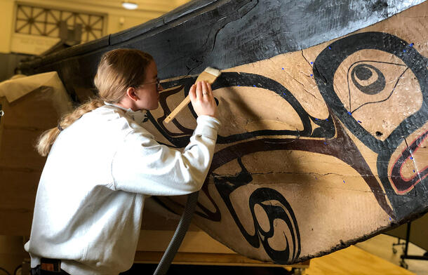Cheyenne Caraway stands leaning towards a canoe mounted on supports, and uses a paint brush to clean a painted area of the canoe,