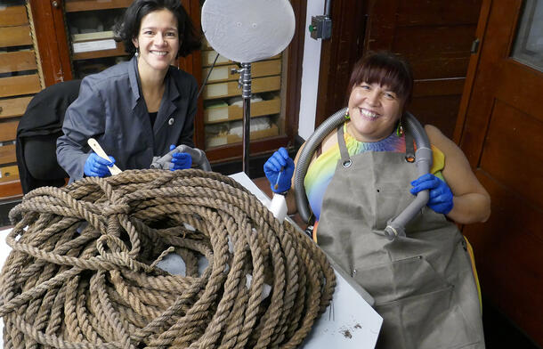 Shyanne Beatty is seated to the right of curator xxx , and wears a protective apron and gloves and holds a hose over a large coil of rope.