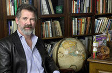 A man in an office looking at the camera with shelves of books and a medium-size globe behind him.