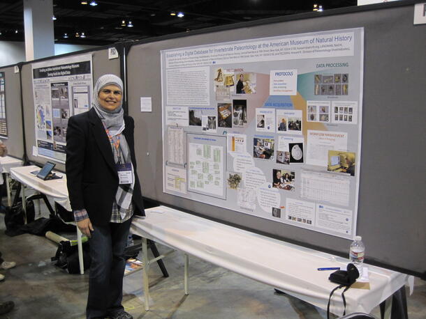 A woman standing beside her research explanation poster in a conference or exhibition hall.