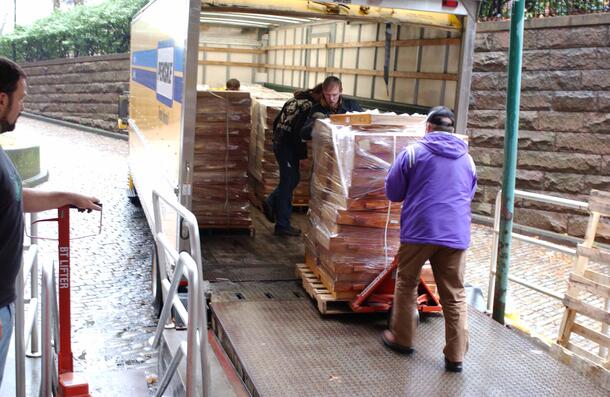 Two men wheeling a pallet down a ramp stacked high with material and wrapped in clear plastic sheeting.