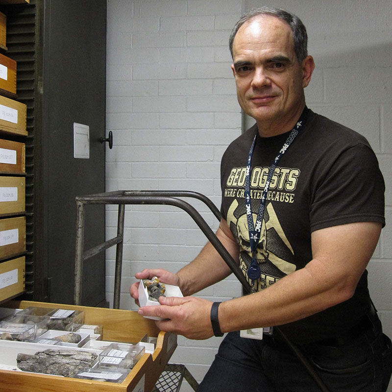 Juan Martinez stands next to a tall shelf with an open drawer that houses small specimens in small open boxes.