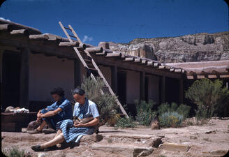 Georgia O'Keefe sits on stone steps and holds a cat in her lap, with a companion seated nearby and an adobe structure in view behind them.
