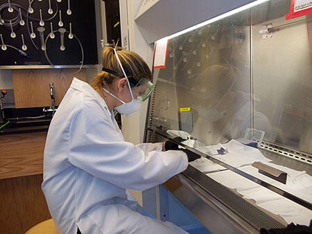 A woman wearing goggles a face mask, gloves, and a lab coat handling something under a laboratory hood.