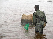 A man dressed in military fatigues standing thigh-deep in water and holding in one hand a small net with a handle.