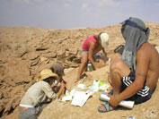 Four people in a dry rocky desert location, three digging, one sitting on the ground with white paper documents at his feet. All wearing head coverings.