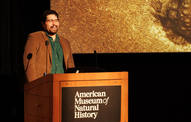 Ernesto Vargas speaks from a lectern, with a partial image of a scientific specimen visible on a large screen in the background.