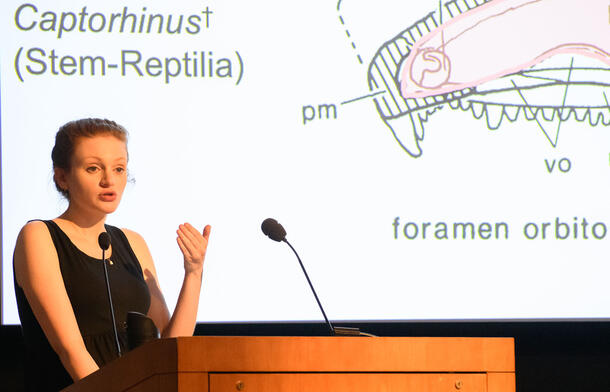 Eva Hoffman speaks from a lectern, with a partial image of Captorhinus visible on a large screen in the background.
