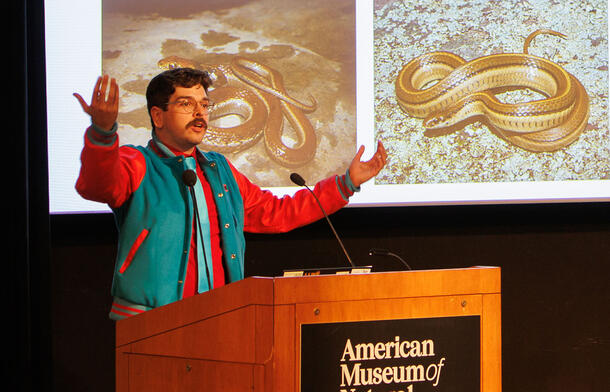 Leroy Núñez speaks from a lectern, with an image of snakes visible on a large screen in the background.
