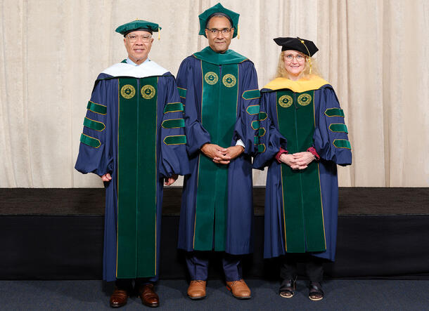Darren Walker, Sean Decatur, and Jo Handelsman wear ceremonial caps and gowns, and stand side-by-side on a platform with a curtain backdrop.