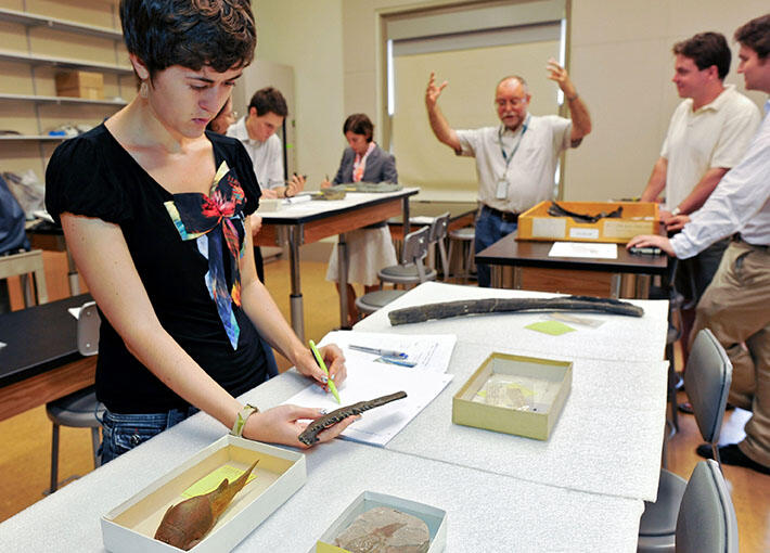 Ph.D. student holds a pencil and a specimen while standing at a lab table, with other students and a professor in the background.