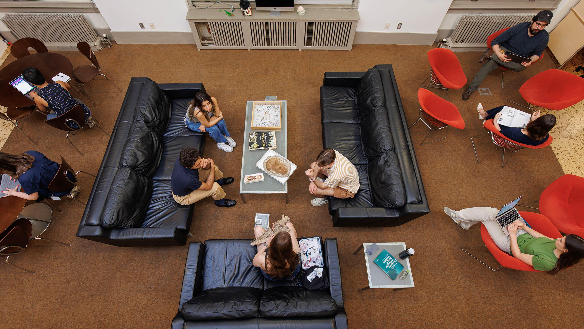 Overhead view of 9 students seated on chairs and couches, conversing and studying in the student lounge for the Richard Gilder Graduate School.