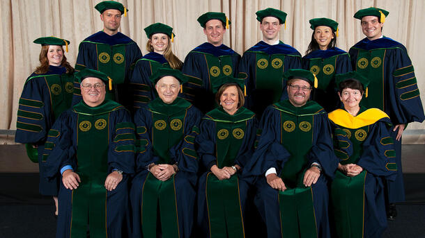 Seven PHD graduates wearing commencement robes stand behind a row of seated Museum mentors and teachers wearing ceremonial robes.