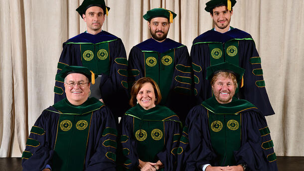 Three PHD graduates wearing commencement robes stand behind a row of seated Museum mentors and teachers wearing ceremonial robes.