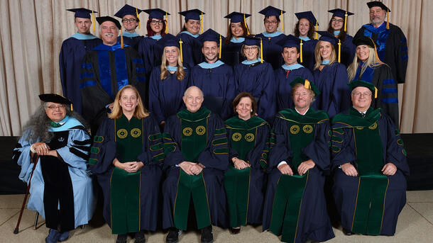 Two rows of MAT graduates wearing commencement robes stand behind a row of seated Museum mentors and teachers wearing ceremonial robes.