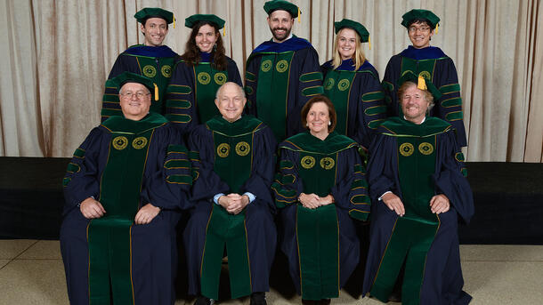 Five PHD graduates wearing commencement robes stand behind a row of seated Museum mentors and teachers wearing ceremonial robes.