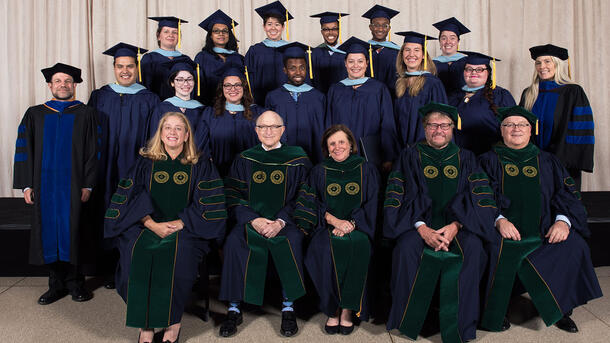 Two rows of MAT graduates wearing commencement robes stand behind a row of seated Museum mentors and teachers wearing ceremonial robes.
