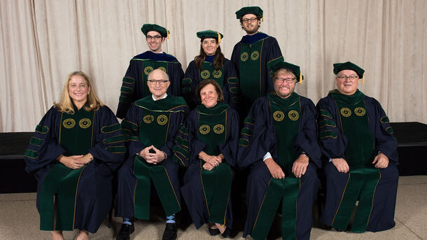 Three PHD graduates wearing commencement robes stand behind a row of seated Museum mentors and teachers wearing ceremonial robes.