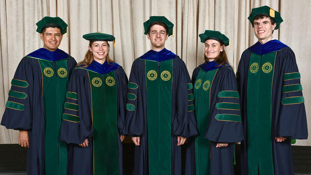 Five PHD graduates stand for a portrait wearing commencement robes.