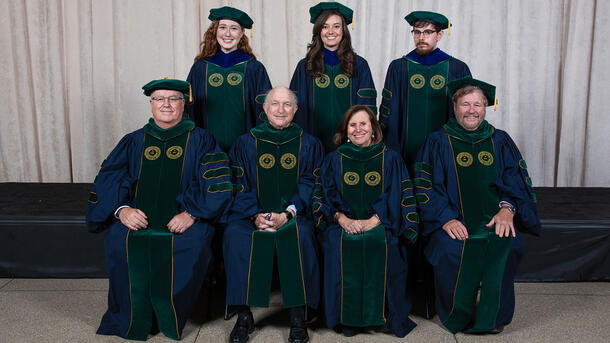 Three PHD graduates wearing commencement robes stand behind a row of seated Museum mentors and teachers wearing ceremonial robes.