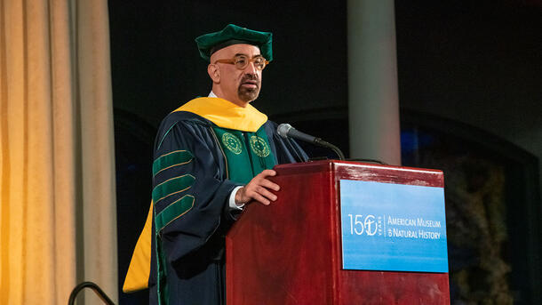 David Spergel wears a ceremonial cap and gown and stands at a podium with a sign reading "American Museum of Natural History.