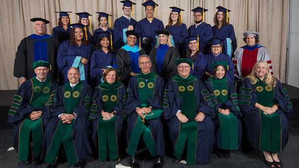 Two rows of MAT graduates wearing commencement robes stand behind a row of seated Museum mentors and teachers wearing ceremonial robes.