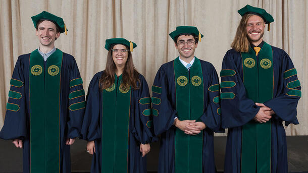 Four PHD graduates stand for a portrait wearing commencement robes.