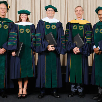 Five adults stand side-by-side wearing ceremonial caps and gowns.