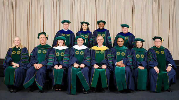 Four PHD graduates wearing commencement robes stand behind a row of seated Museum mentors and teachers wearing ceremonial robes.
