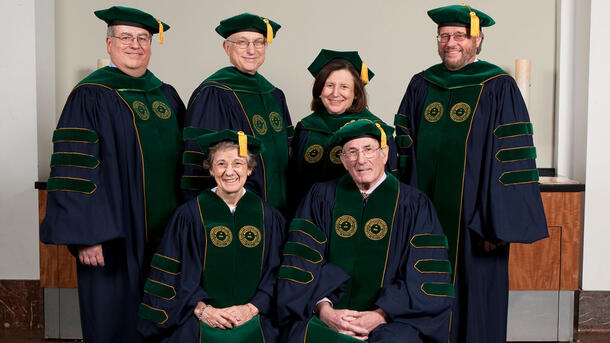 Two honorary degree recipients wearing ceremonial robes and are seated in front of four standing Museum representatives wearing ceremonial robes.