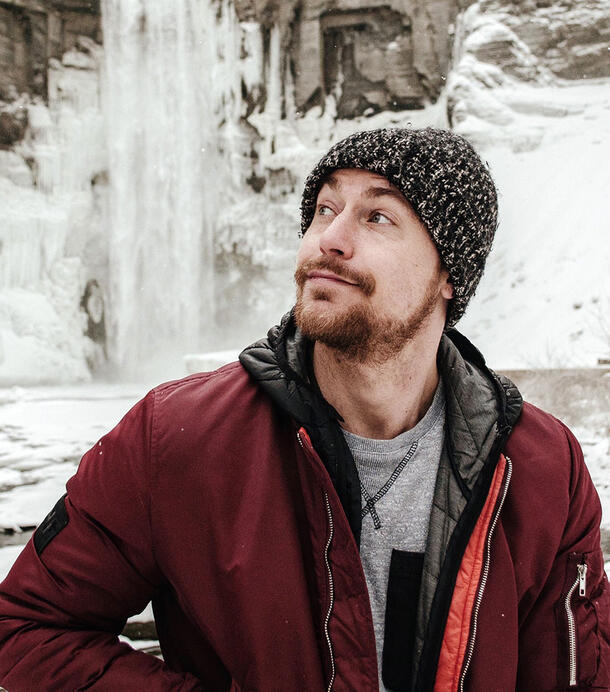 Daniel Hooper wears a warm weather hat and jacket and stands in front of the icicles of a frozen waterfall.
