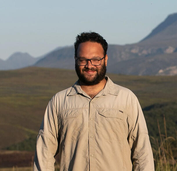 Pedro H. N. Bragança poses for a smiling portrait in the field, with a mountain range in view behind him.