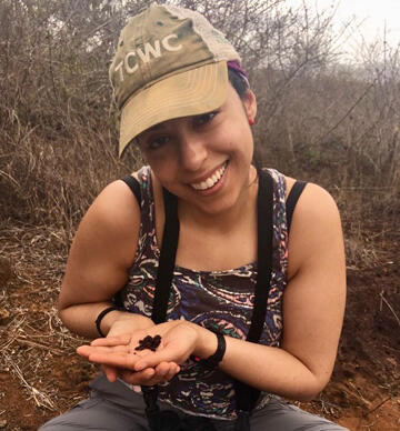 Johanna Harvey wears a baseball cap, and is seated outside in a brushy area and holding a small specimen in her hands.