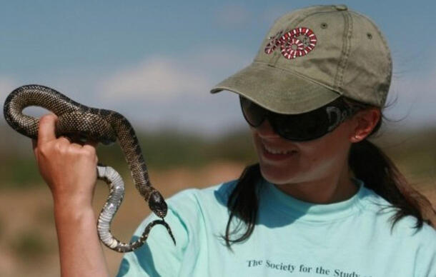 Sara Ruane holds a snake in one hand while wearing sunglasses and a baseball hat with a snake image on it. 