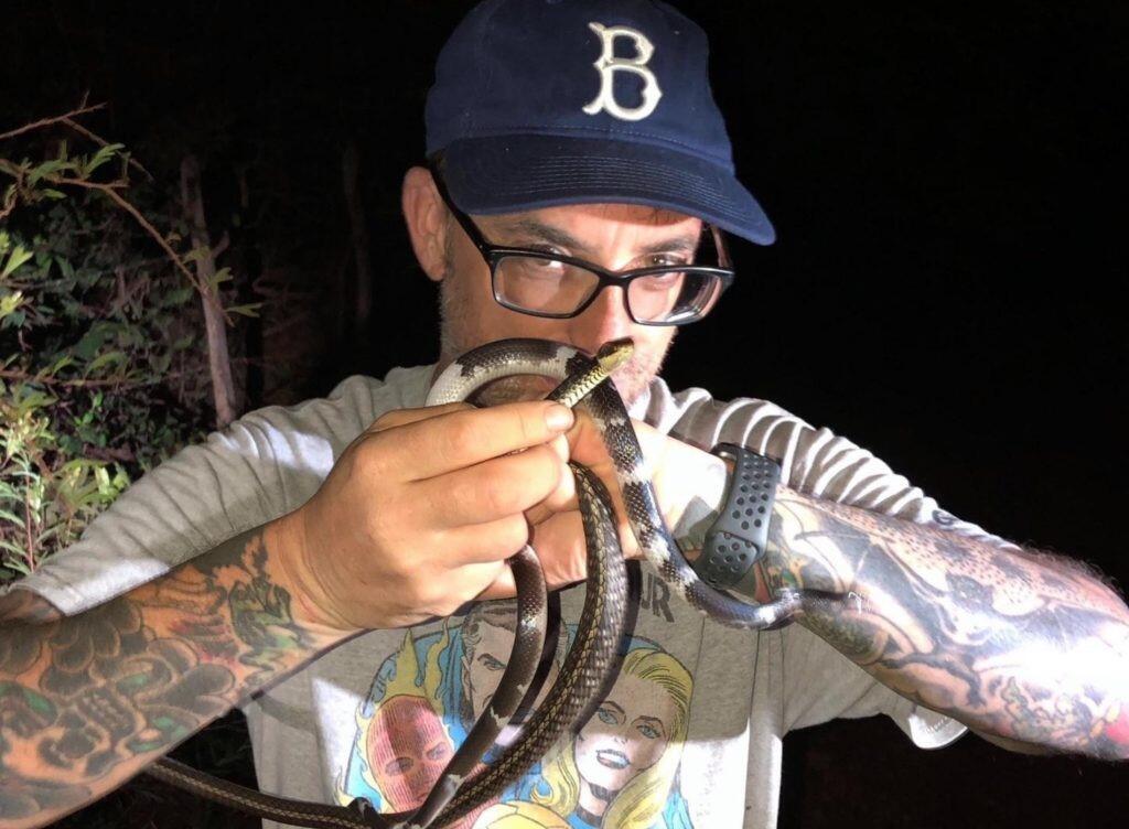 Frank T. Burbrink wearing a baseball cap and holding a snake up, obscuring his face slightly.