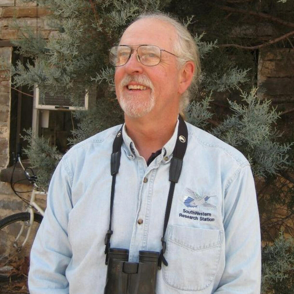 Bearded man with white hair, smiling, looking up, wearing binoculars.