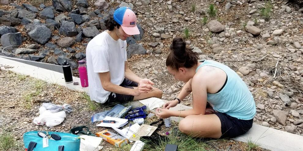 Young man and woman sitting on the ground with a variety of supplies for their field work surrounding them.