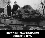 A black and white photo of two men atop a flat bed vehicle bearing a massive rock with a pitted surface. The caption superimposed at the bottom of the photo states: "The Willamette Meteorite comes to N Y C."