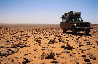 A sports utility vehicle with equipment on its roof rack parked on a sandy desert that is ittered with small and medium-size stones.