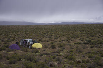 A panoramic view of a wide flat land area with scrubby plants and two people near a parked van, with two small domelike tents set up on the ground nearby.