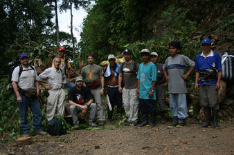 Eleven men posing for the camera in a clearing with green vegetation in the background, dressed in T- shirts, trousers, baseball caps, and rubber boots.