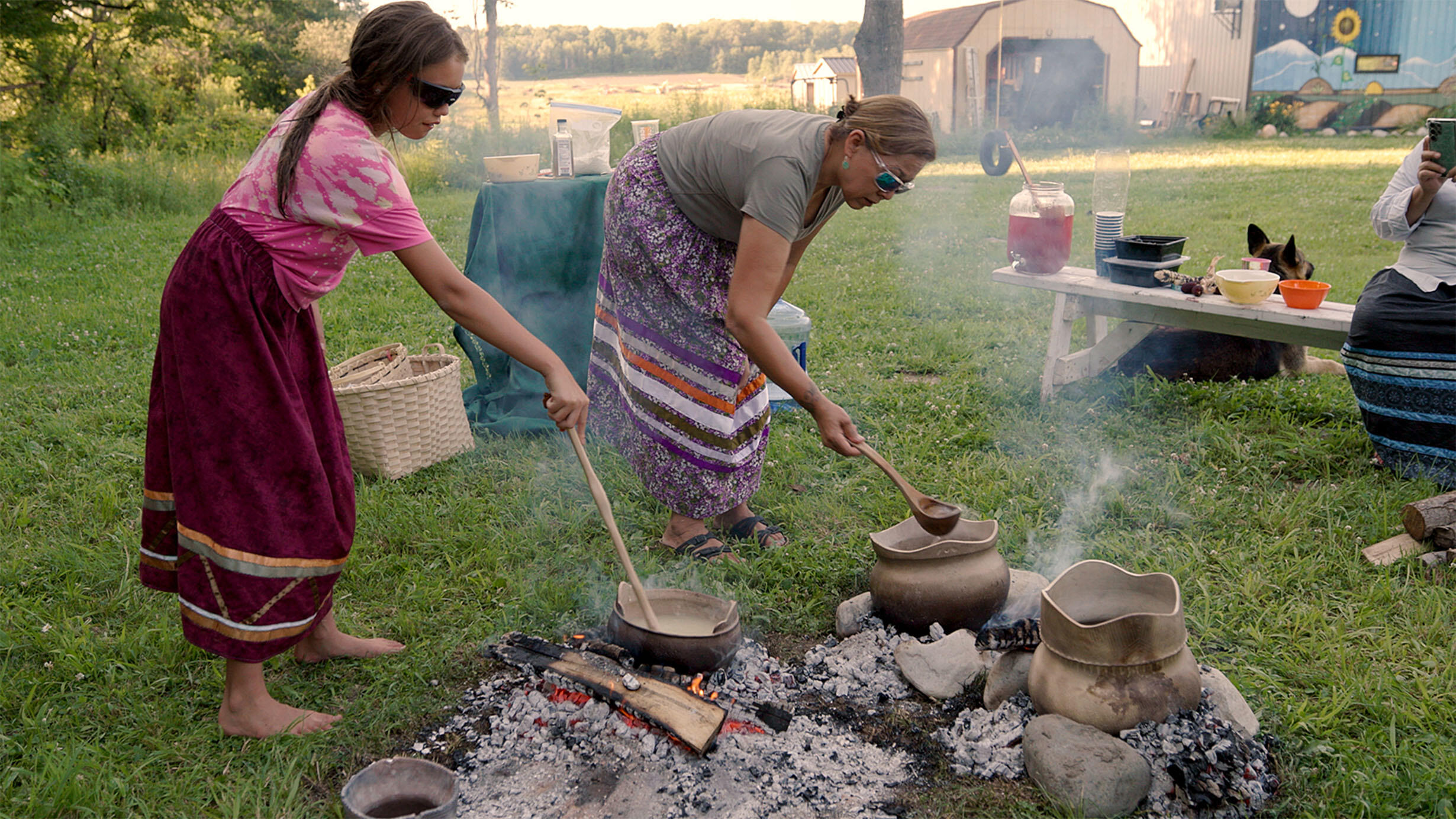 Two people stand outside and use ladles to stir food cooking in clay pots on a fire.