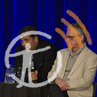 Neil deGrasse Tyson leans on a table and speaks into a microphone, Ross MacPhee sits beside him, listening.